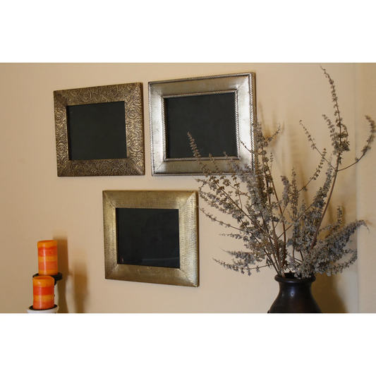 Three decorative, ornate frames on a beige wall, accompanied by a vase with dried plants and two candles. Wood frames set. Brass clad mango wood.