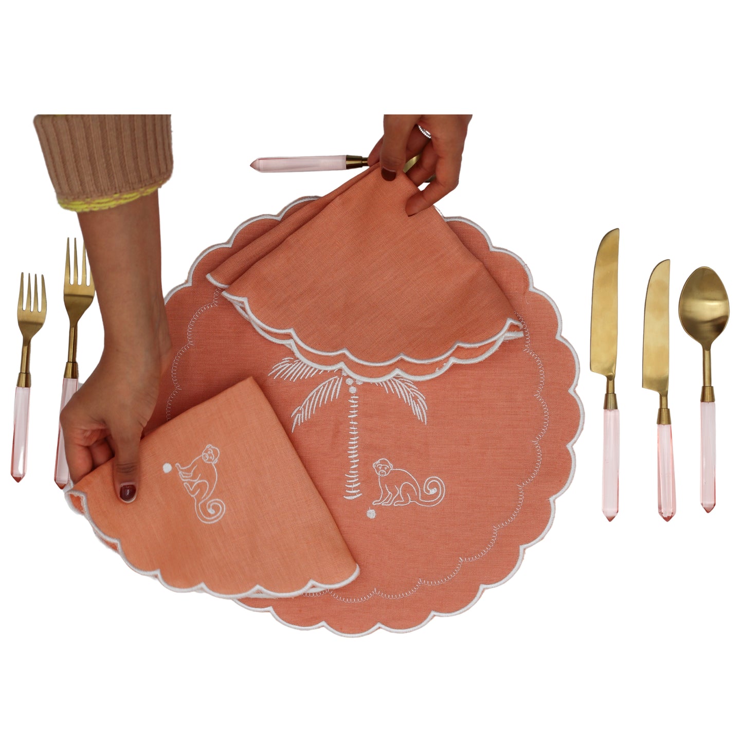 Coral placemats with white designs held by a hand, surrounded by gold and pink cutlery on a white background.