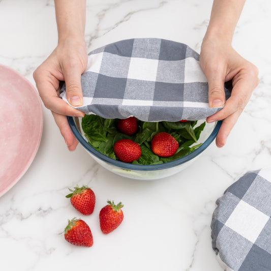 Person holding a bowl with a checkered cover, surrounded by strawberries on a marble surface. Checkered cloth covering a bow. Elastic for proper fit. Flax Linen.
