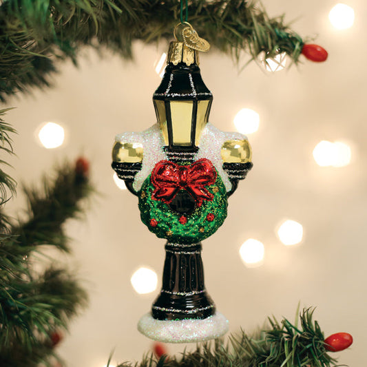 A decorative Christmas ornament shaped like a black street lamp with gold accents, a green wreath with a red bow, and white snow-like detailing, hanging on a Christmas tree branch with warm blurred lights in the background.