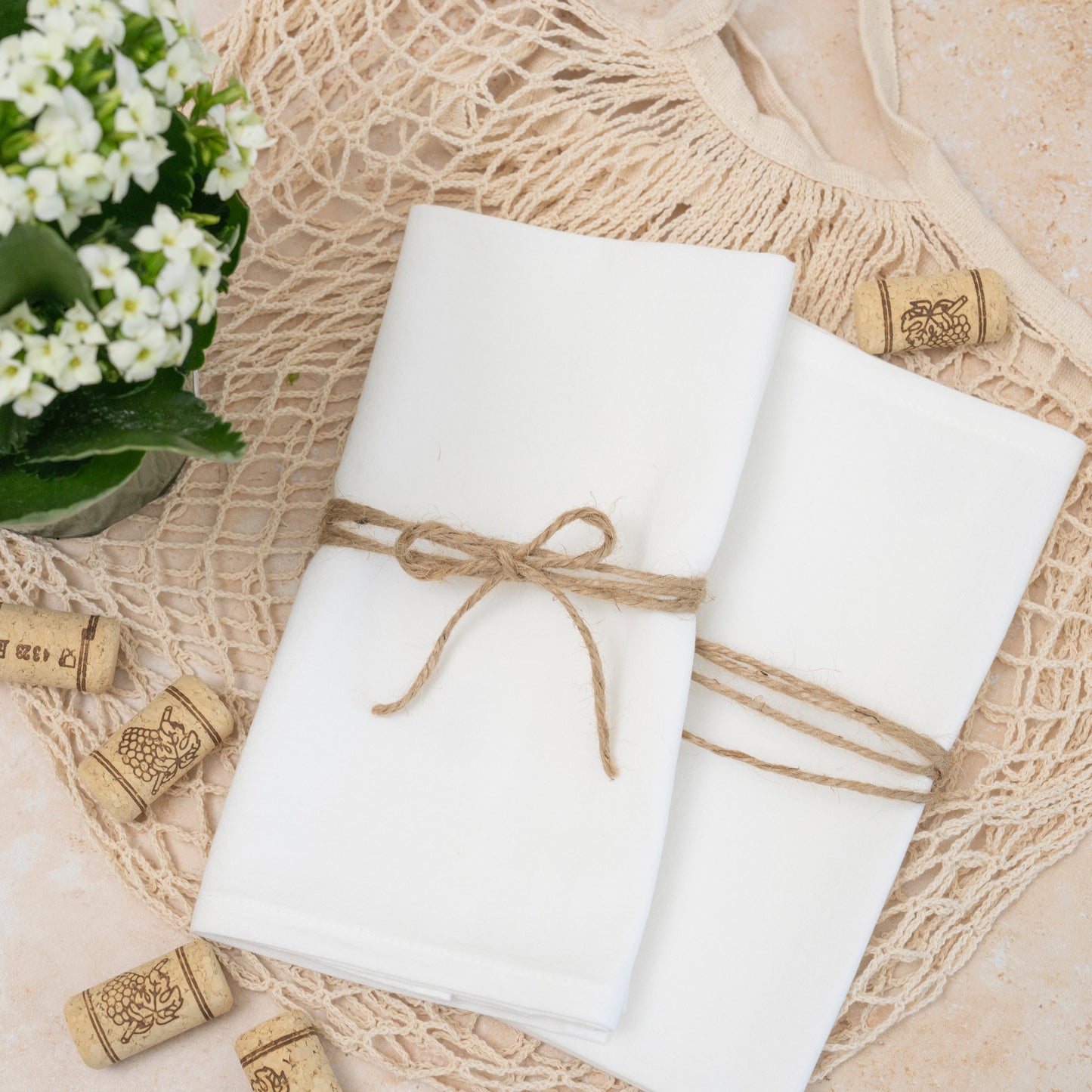 Two white wrapped gifts tied with brown string on a textured surface with wine corks and flowers.