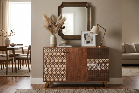 A mid-century modern wooden sideboard cabinet in a warm medium brown finish, featuring patterned woven and geometric wood panels on the doors and drawers. The cabinet is styled in a living room setting with a matching wood-framed mirror, a table lamp, a vase of pampas grass, and framed art.