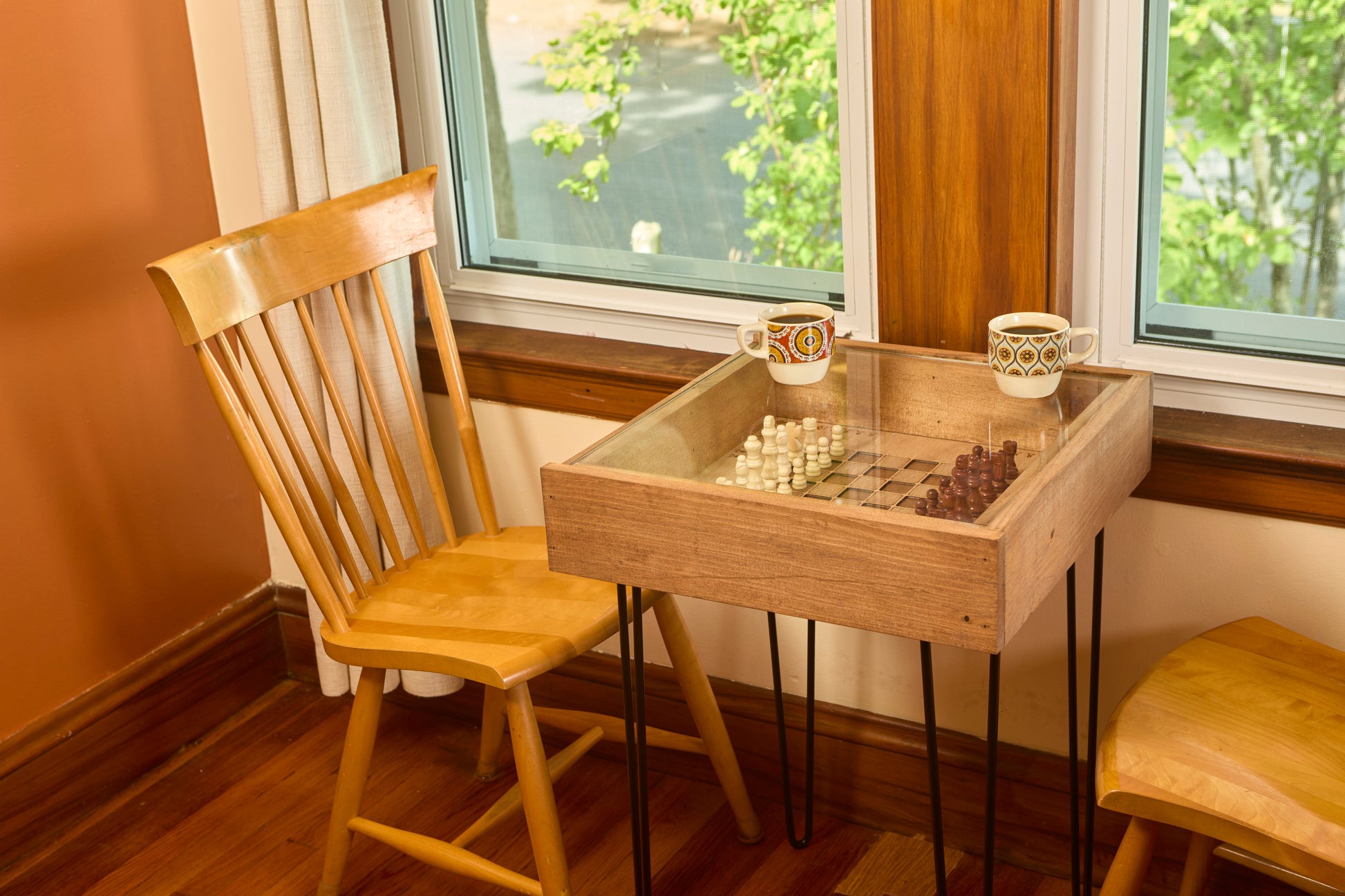 Wooden table with glass top and chess pieces next to a chair in a room with windows.