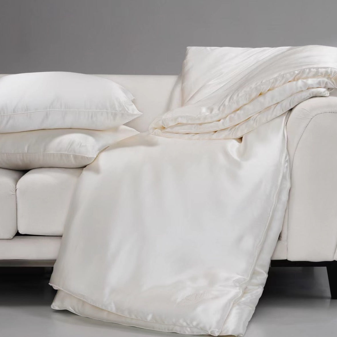 White silk comforter and pillows on a gray sofa against a gray background