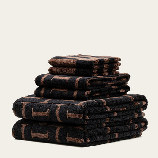 A stack of folded bath towels in a dark brown and black striped pattern, set against a solid off-white background.