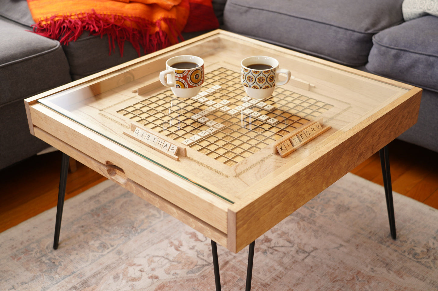 Wooden coffee table with a scrabble board and two cups in a living room setting.
