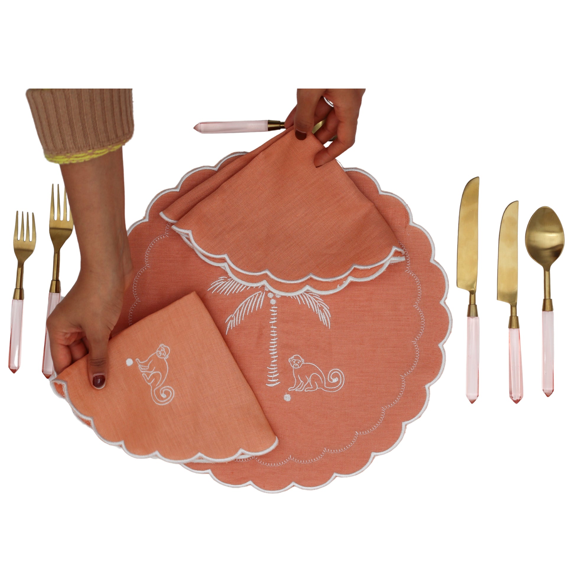 Coral placemats with white designs held by a hand, surrounded by gold and pink cutlery on a white background.