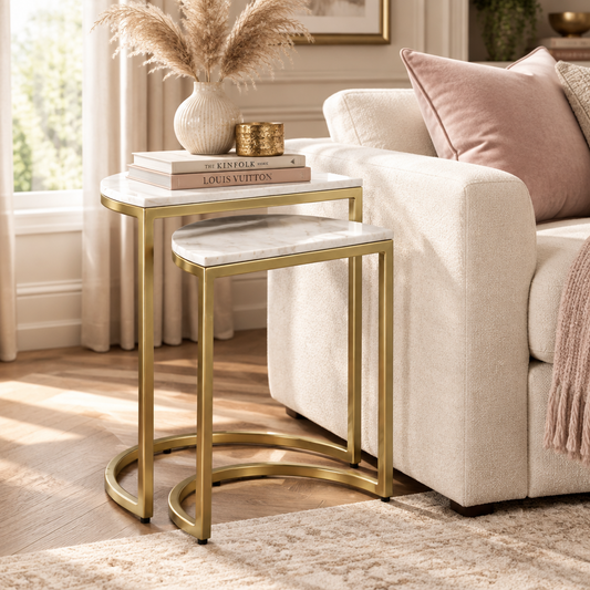 A gold metal side table with a white marble top and matching marble-topped stool in a bright living room beside a cream sofa. Decorative books, a vase with dried grass, and small gold candle holders sit on the table.