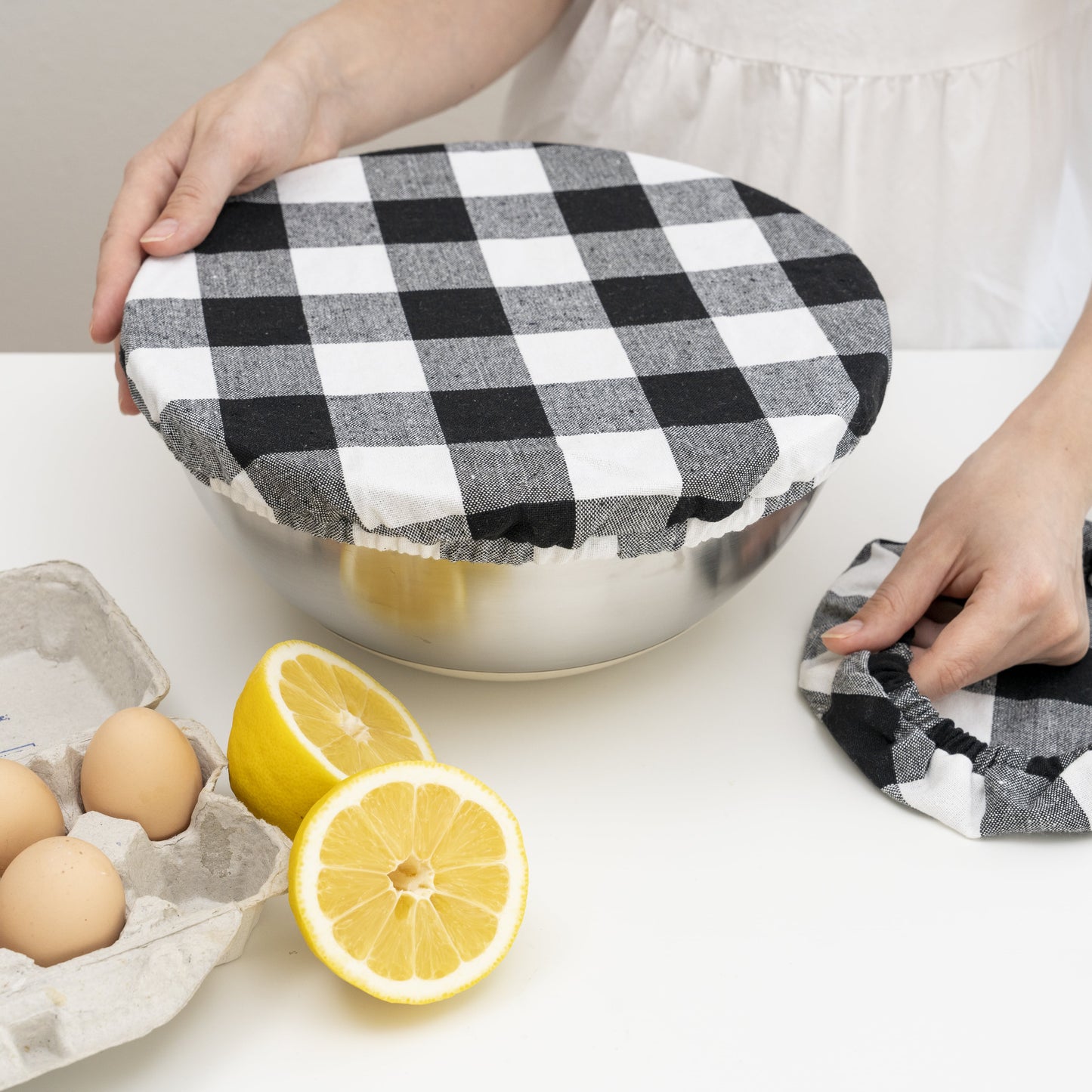 Person using a black and white checkered cloth to cover a metal bowl on a kitchen counter with lemons and eggs. Plaid cloth covering a bowl.  Elastic for proper fit. Flax Linen.