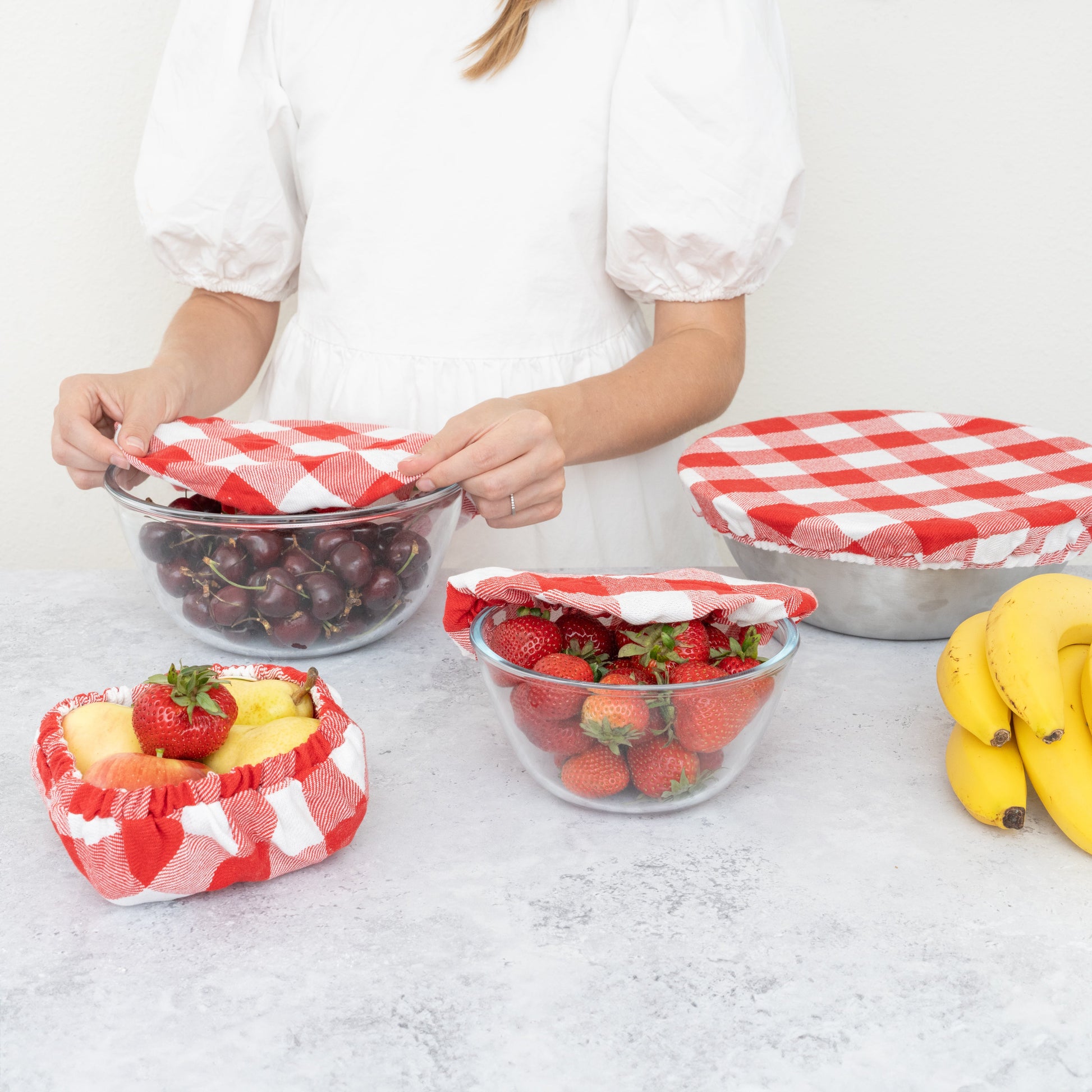 Person arranging fruits in glass bowls with red and white checkered covers on a light surface. Plaid cloth covering a bowl.  Elastic edge for proper fit. Flax Linen.