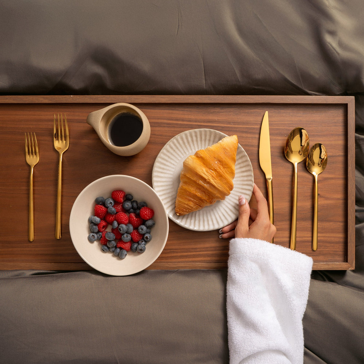 A top-down view of a wooden breakfast tray set on a bed, featuring a croissant on a striped plate, a bowl of fresh berries, a small creamer, and gold-toned cutlery. A person's hand in a white robe is reaching toward the croissant.