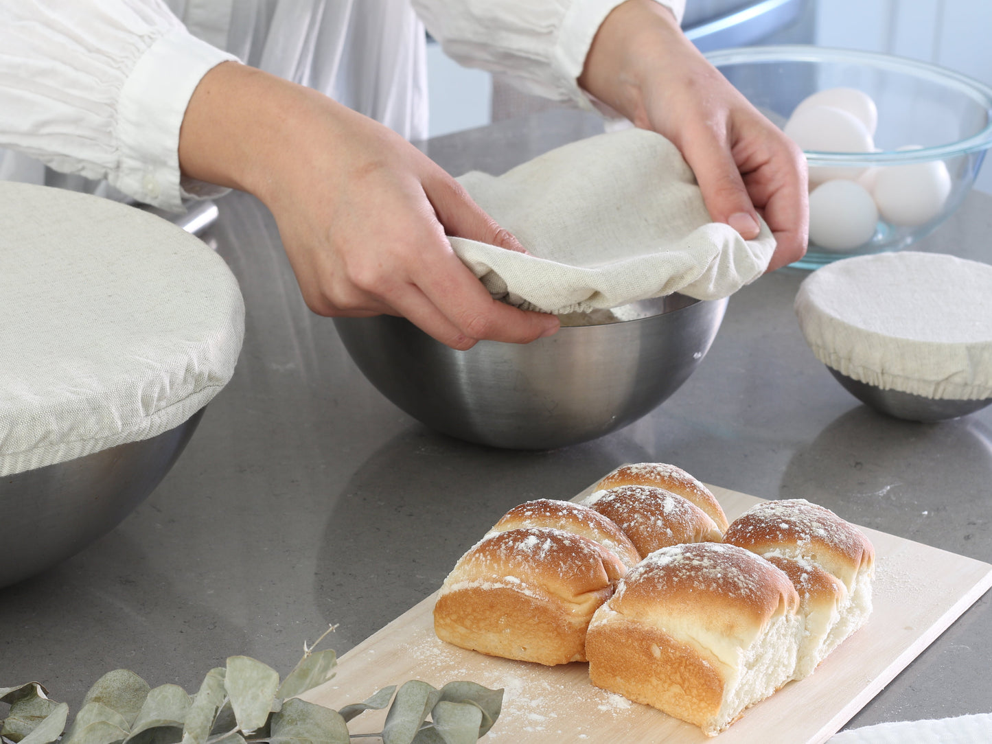 Baked bread rolls on a cutting board with a person preparing ingredients in the background. Wheat/beige cloth covering a bowl.  Elastic for proper fit. Flax Linen.