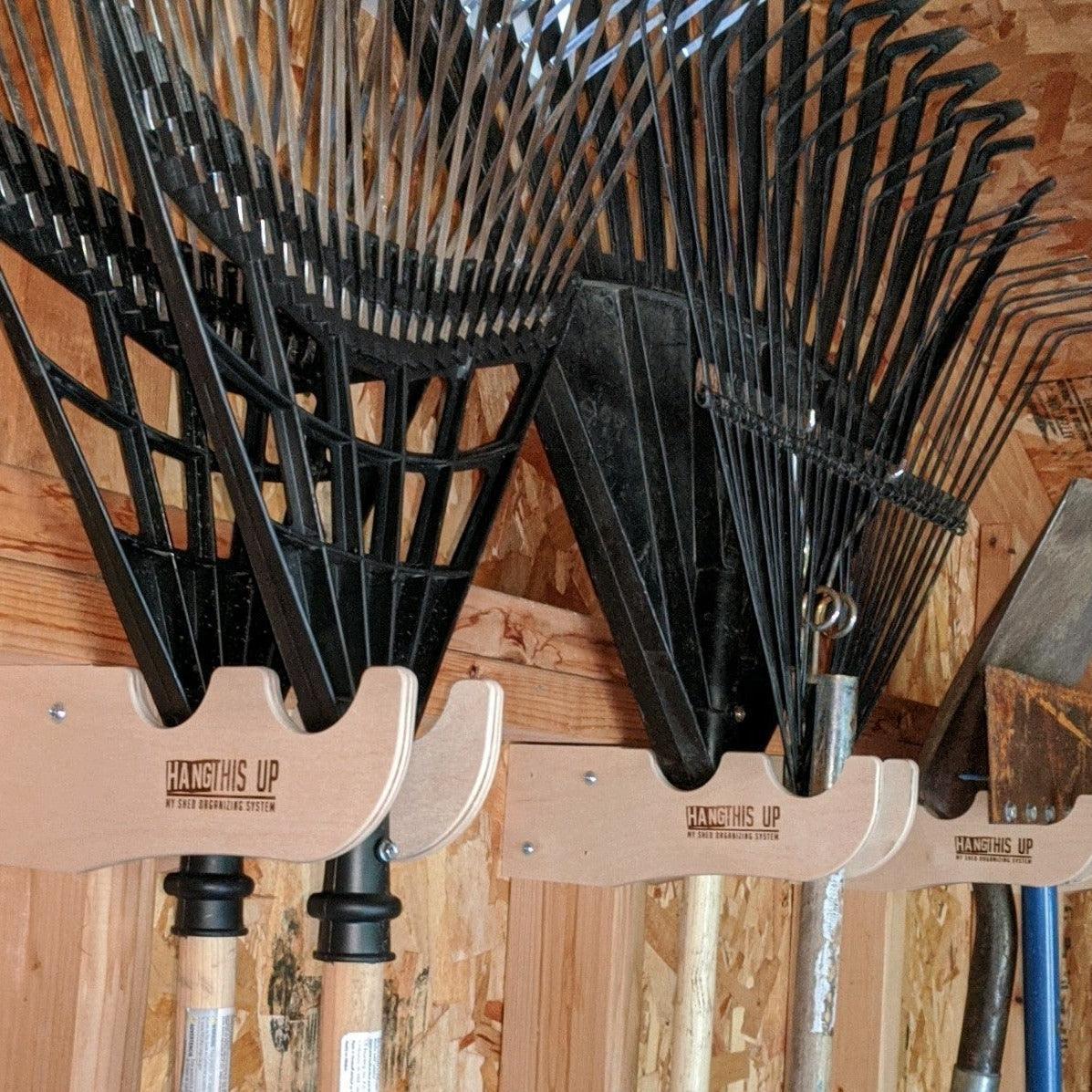 Set of black garden rakes hanging on a wooden wall with 'Hang This Up' tool holder.