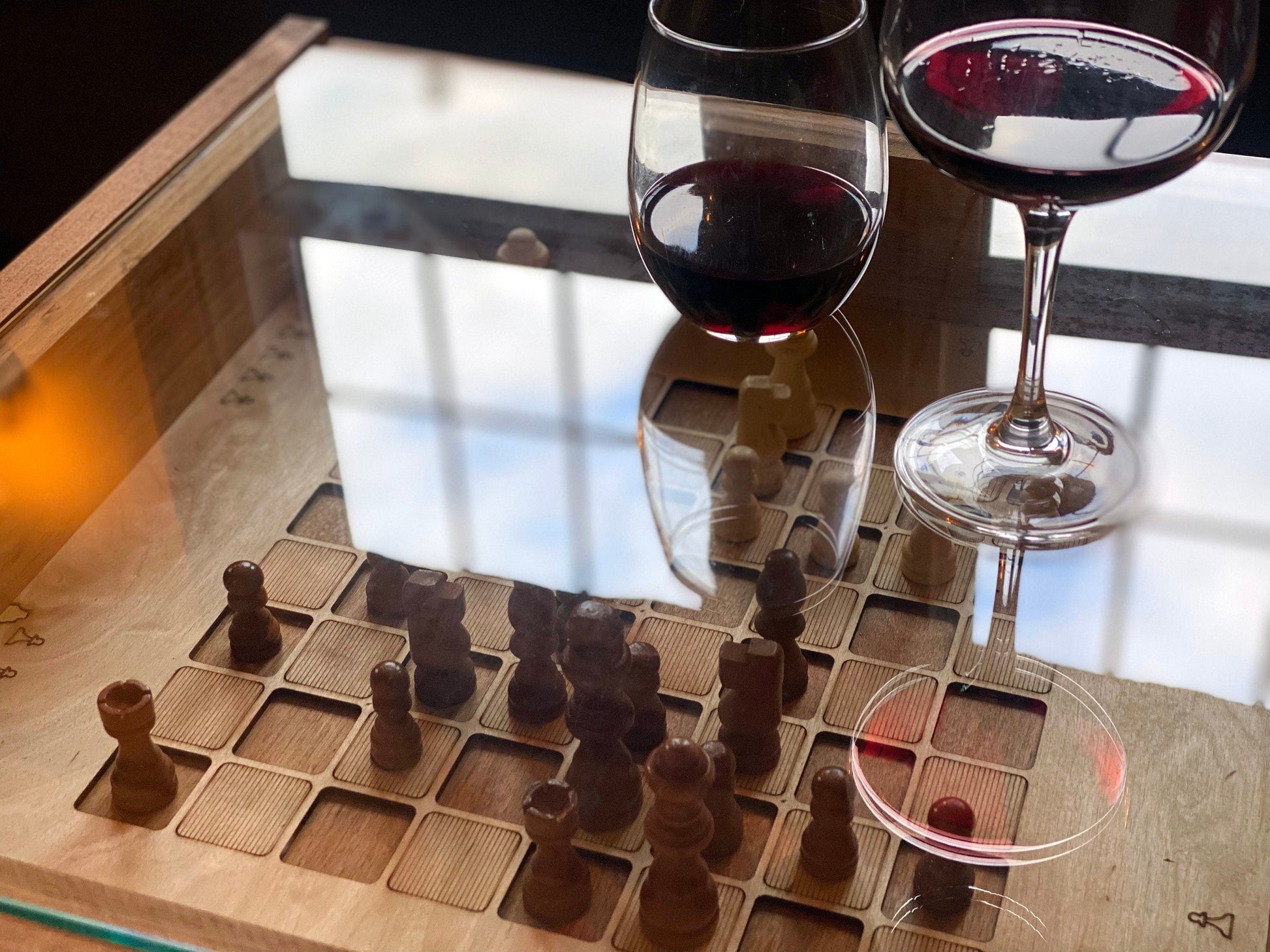 Two wine glasses filled with red wine on a chessboard with wooden pieces.