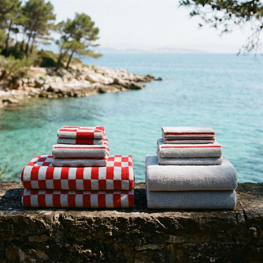 Stacked folded towels displayed outdoors on a stone ledge with a coastal sea and trees in the background. One stack features a red and white checkered pattern, and the other is solid light gray.