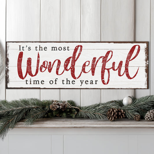 A rectangular white wooden sign with a dark rustic border reads “It’s the most Wonderful time of the year” in red and black lettering, displayed on a mantel with a green pine garland and pinecones against a white paneled wall.