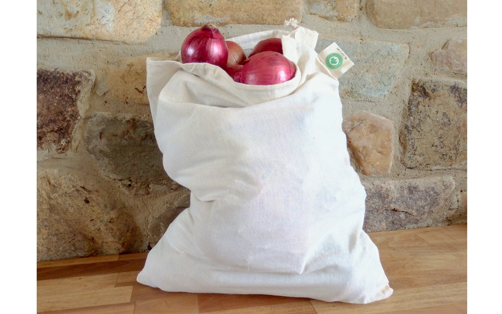 White reusable shopping bag with red onions against a stone wall background. 100% Cotton Produce Bags.