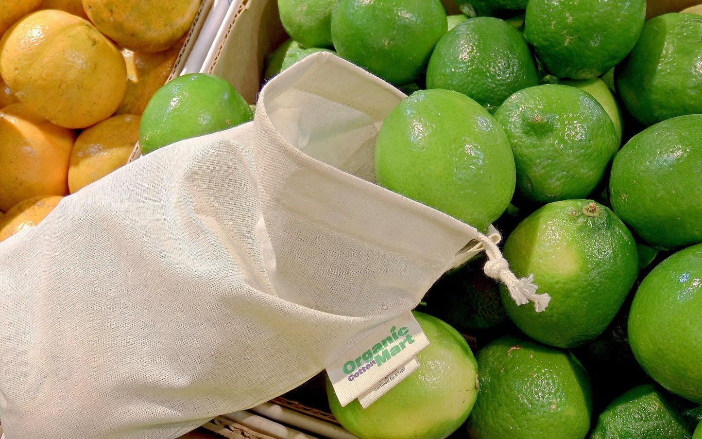 Green limes in a cloth bag with an 'Organic' label, surrounded by oranges. 100% Cotton Produce Bags.