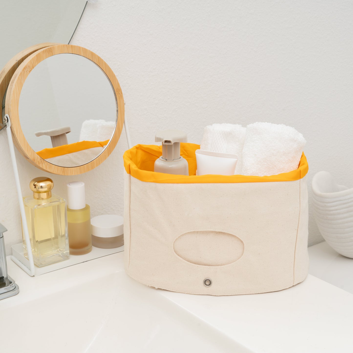 Bathroom scene with a beige storage basket with an orange lid containing towels and toiletries, next to a round mirror.